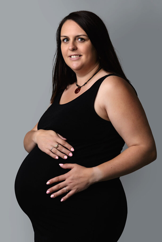 Pregnant woman standing with hands resting on her bump, photographed in neutral light.
