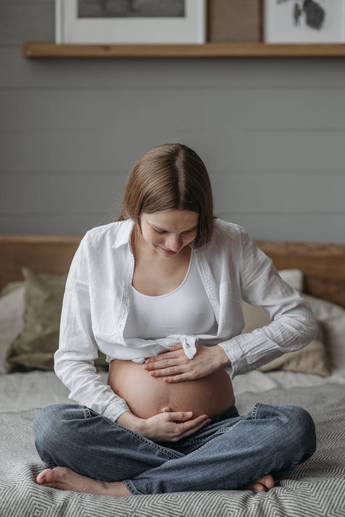 A serene portrait of a pregnant woman sitting on a bed, gently holding her baby bump.