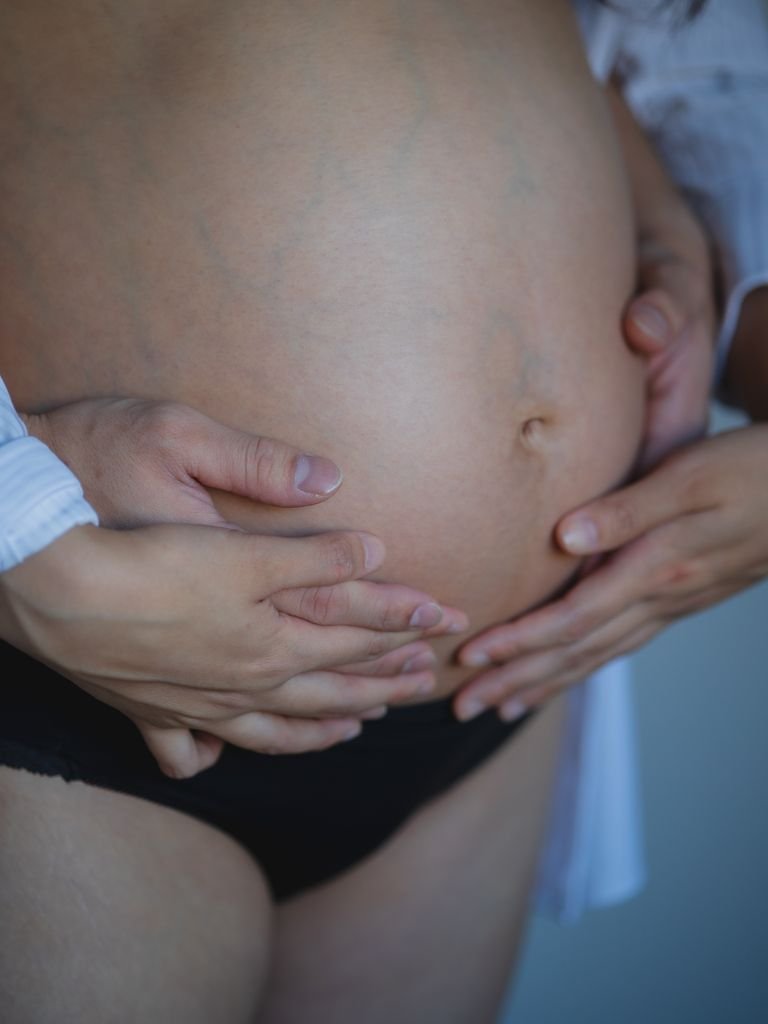 Faceless pregnant woman wearing black underwear and shirt touching naked belly with anonymous man while standing in room at home